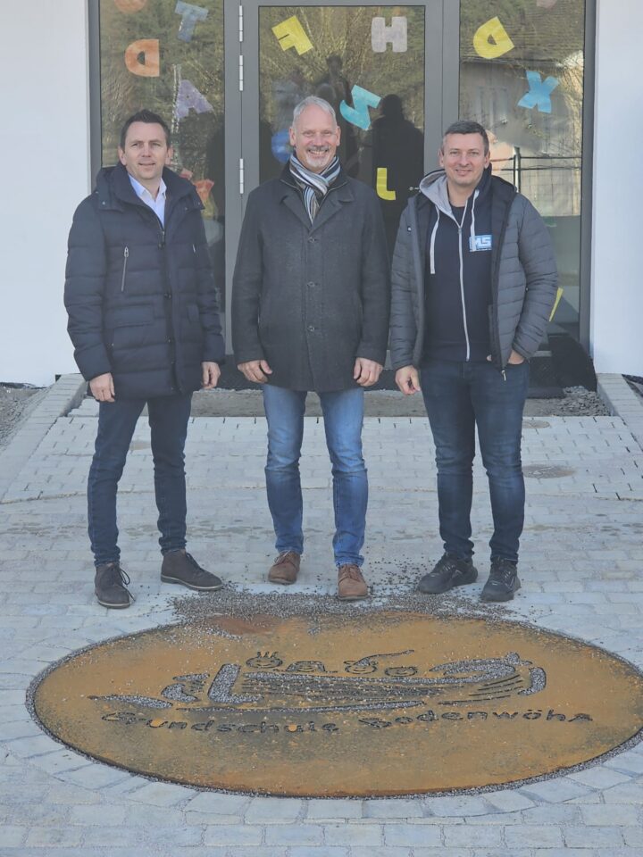 Mathias Schießl (MS Metalltechnik GmbH), Bürgermeister Georg Hoffmann und Paul Michalski (MS Metalltechnik GmbH) stehen vor dem Haupteingang der neuen Grundschule Bodenwöhr hinter der gespendeten Boden-Logotafel aus Metall.